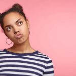 Dreamy funny stylish female with African appearance pouts lips, looks aside, dreams about something pleasant, wears earrings and casual striped sweater, poses against pink wall with copy space