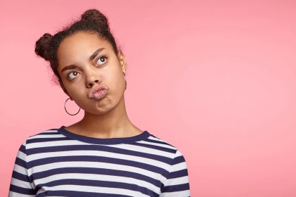 Dreamy funny stylish female with African appearance pouts lips, looks aside, dreams about something pleasant, wears earrings and casual striped sweater, poses against pink wall with copy space