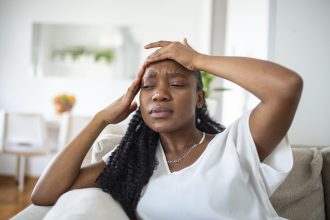 Portrait of a young black girl sitting on the couch at home with a headache and pain. Beautiful woman suffering from chronic daily headaches. Sad woman holding her head because sinus pain