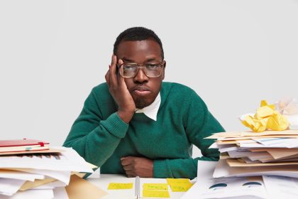 Bored male worker has tired facial expression, looks in displeasure, works long hours on project task, has dark healthy skin, sticks notes in notepad, tries not to forget everything, isolated